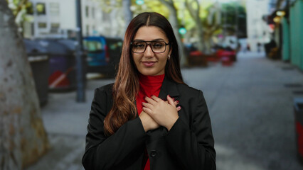 Fototapeta premium Woman smiling with eyes closed and hands on heart, standing on a vibrant city street wearing a red top and black jacket, exuding warmth and confidence.