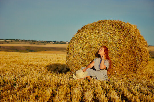 red-haired beautiful woman with hat in field with haystacks against blue sky