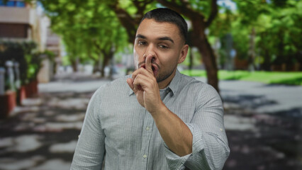Man bringing finger to lips for silence on city street under leafy trees in bright sunlight;...