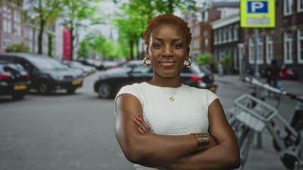 Woman in white top folds arms on city street lined with parked cars; confidence empowerment...