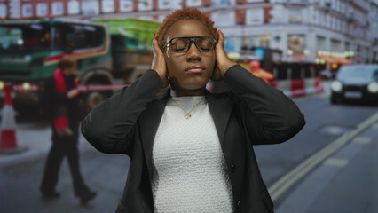 Woman wearing glasses and blazer covers ears on busy urban street amid blurred traffic; sensory overload.