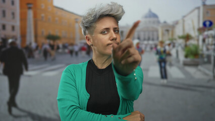 Woman with short hair and green cardigan points finger on street amid crowds in front of baroque...