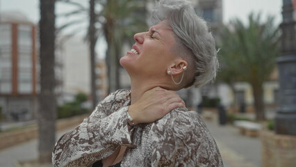 Caucasian woman in a patterned blouse holding neck in busy city street lined with buildings;...