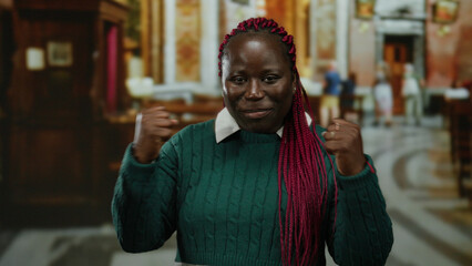 Woman expressing excitement indoors in a church setting with vibrant hair and green sweater, capturing joy and emotion amidst blurred architectural background.