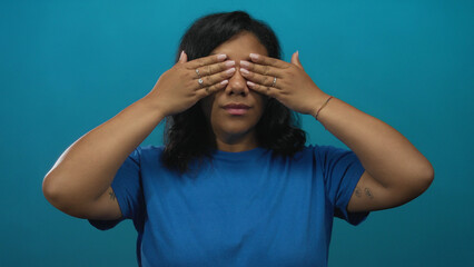 Dark skinned african american woman covers eyes with hands and smiles subtly in studio on blue...