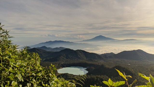 A breathtaking sunrise from Mount Sagara in Garut, overlooking the serene Talaga Bodas crater lake. The soft morning light paints the sky in warm colors, while mist rises gently over the mountains, cr