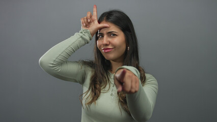 Young hispanic woman making loser gesture with hand on forehead against a grey wall in studio...
