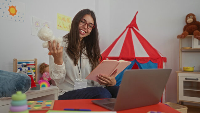Hispanic woman teacher reading to preschool children in a vibrant kindergarten setting with toys tent and laptop