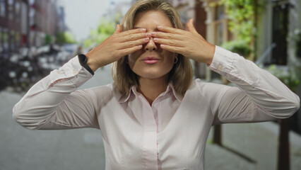 Fototapeta premium Woman with blonde hair in pink shirt covers eyes with hands on city street in daylight; joy playful excitement.