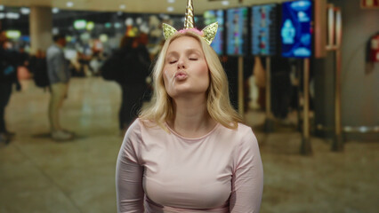 Blonde woman in unicorn diadem playfully poses at airport terminal, adding humor to the bustling...