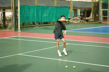 Asian male tennis player jumps to hit a powerful forehand shot on a sunny outdoor court during a practice session