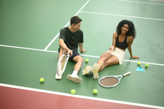 African female tennis player and Asian male friend sitting on court smiling and chatting after a tennis match, surrounded by rackets and balls