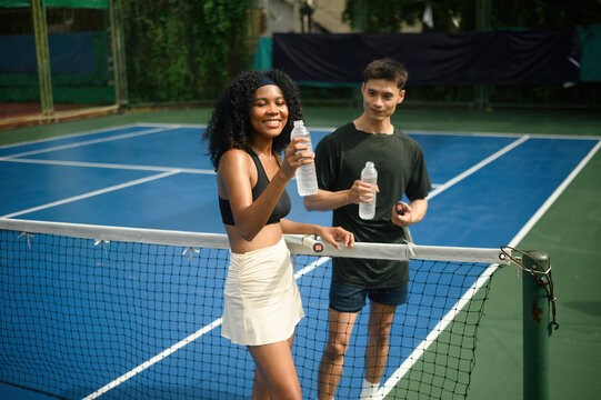 Happy mixed-ethnicity tennis partners enjoy a refreshing drink together, staying hydrated after an intense game