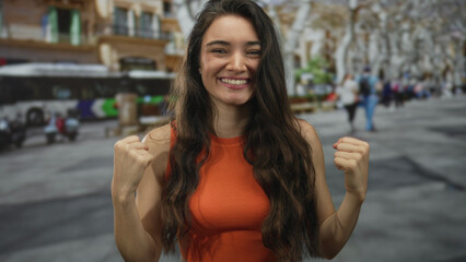 Young hispanic woman in orange top flexes bicep on busy sunlit city street corner beside bus; confidence.