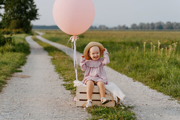 Happy little girl sits alone on road in middle of field holding large pink balloon in summer. Portrait of cute smiling child playing in nature. Children's birthday party or outdoor nature photoshoot.