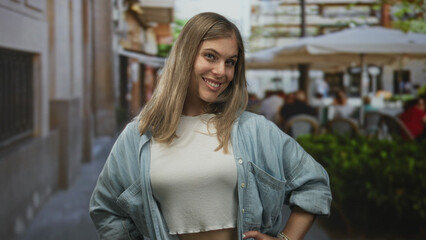 Fototapeta premium Woman stands with hands on hips and smiles beside outdoor restaurant tables on street; carefree confidence.