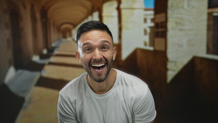 Young hispanic man smiling on university campus with old architecture in the background suggesting a lively academic environment.