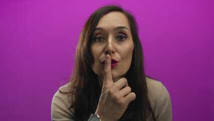 Fototapeta premium Woman gesturing silence against vibrant pink background with serious expression and long brown hair suggesting secrecy and calm.