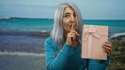 Senior woman with grey hair gesturing silence while holding a pink gift box at the seaside beach setting with ocean backdrop under a serene sky outdoors.