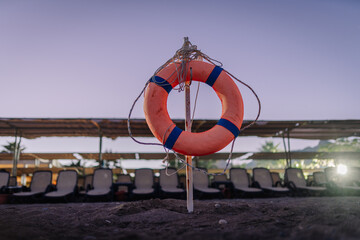 A lifebuoy ring mounted on a pole in the sand at a beach with empty loungers and canopies in the background during dusk.