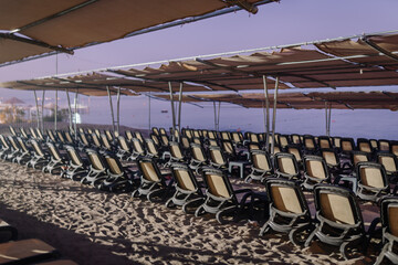 Rows of empty beach loungers under shade canopies on a sandy shore, facing a calm sea during early morning or evening light.