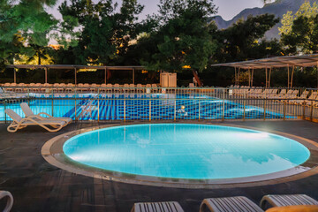 Illuminated swimming pool area at night with lounge chairs, surrounded by trees and mountains, creating a peaceful vacation setting.