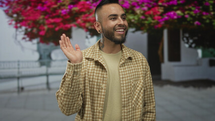 Young man smiling and waving his hand on a sunny street by white building with pink bougainvillea overhead; casual warmth.