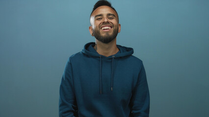 Man smiling showing teeth and beard in teal studio wearing blue hoodie, relaxed pose; joy warmth openness.