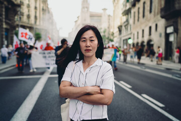 Fototapeta premium Asian woman confidently faces camera with arms folded, holding phone, representing digital independence, presence and tech-driven strength in public context.
