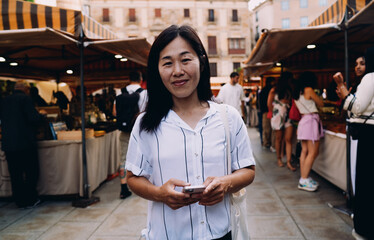 Portrait of smiling Asian woman at street market holding smartphone, standing confidently in urban crowd, reflecting real-time connection and cultural openness.