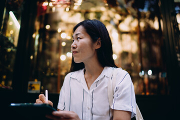 Asian woman with digital tablet looks away, pausing from creative process, reflecting calm focus and independent thinking in ambient urban setting.