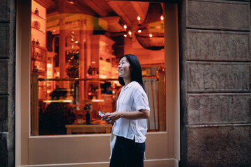 Asian woman stands outside warmly lit cafe, holding phone in hand, embracing peaceful digital pause during an urban stroll and reflecting quiet joy in everyday connectivity.