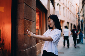 Young Asian woman takes selfie beside warm-lit shop window, using smartphone to capture vibrant moment while exploring city, merging travel, tech, and personal branding.
