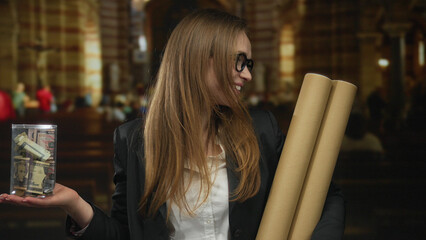 Woman holds transparent donation jar filled with banknotes and cardboard tubes in church interior; generosity.