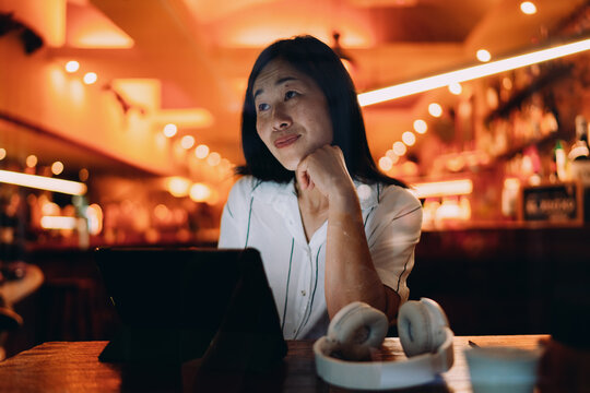 Contemplative Asian woman reflects while seated with digital tablet, portraying slow tech use, intentional browsing, and mindful presence in softly lit modern space.
