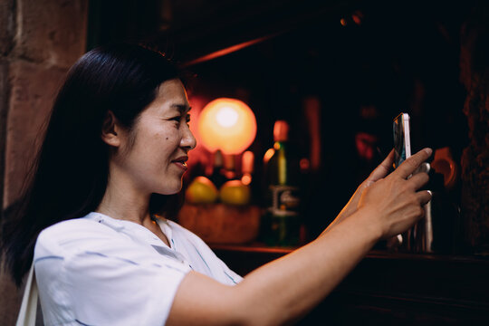 Asian woman takes smartphone photo at bar counter, enjoying solo exploration and capturing detail with mobile technology in low-light intimate environment.