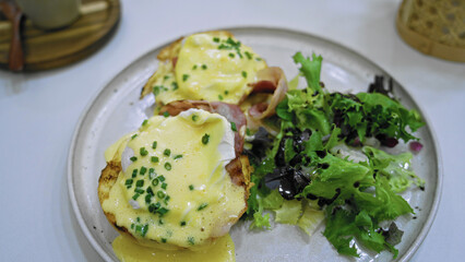 Eggs benedict with creamy hollandaise sauce and fresh salad served on a white plate in a cafeteria setting showcasing a delicious breakfast meal.