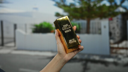 Man holding a shiny gold bar on a street outdoors in a city setting, emphasizing wealth and...