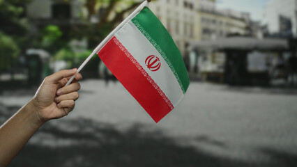Hand holding iranian flag outside on city street with blurred background, signifying patriotism,...