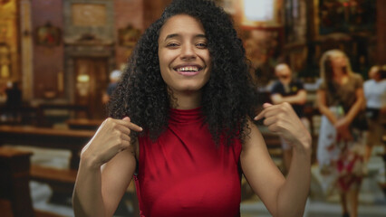 Young hispanic woman with curly hair in red top points finger to herself in a church building; self...