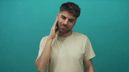Young hispanic man wearing beige t shirt cups cheek for toothache in teal studio setting;...