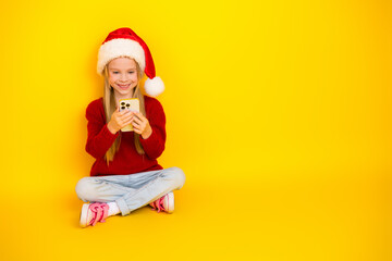 Playful Christmas Kid Seated in Santa Hat Texting on a Bright Yellow Studio Backdrop for Marketing and Social Media Use in cool seasonal season creative advertising campaigns across digital platforms