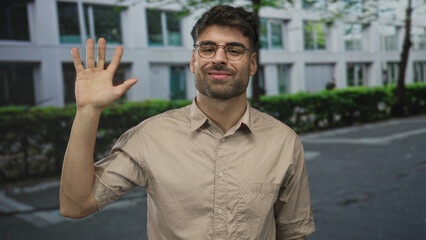 Naklejka premium Man in beige shirt and glasses raising hand in greeting on sunlit tree lined city street; friendly greeting.