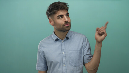 Man looking upward while pointing finger in studio against teal wall wearing light blue shirt;...