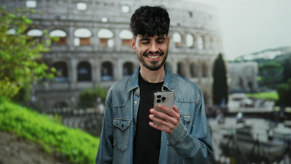 Young hispanic man smiling while holding smartphone near the roman coliseum, showcasing an outdoor...