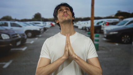 Man in white t shirt stands with pressed hands in prayer on street amid rows of parked vehicles...
