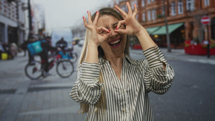 Fototapeta premium Blonde woman wearing glasses makes ok sign with fingers near eyes on downtown busy city street; playfulness.