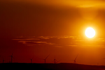 sunset over a wind farm with wind turbines silhouetted against the orange sky. A scenic landscape symbolizing renewable energy, sustainability, and clean power from nature.”