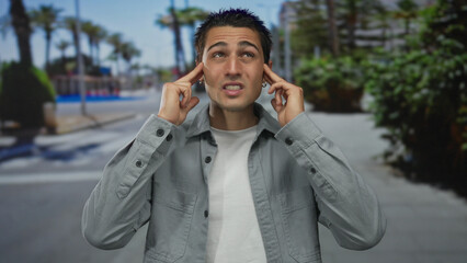 Young man concentrating with fingers on temples standing on urban street with palm trees in background, wearing casual gray shirt in vibrant city environment.