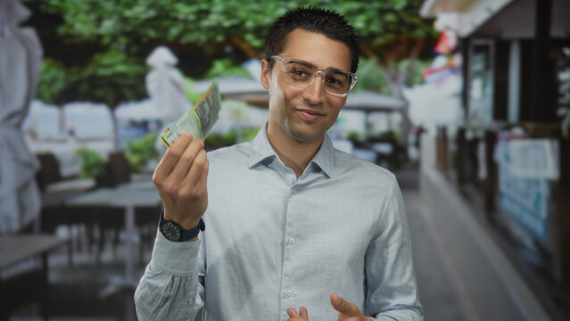 Young man holding australian dollars in a relaxed outdoor cafe terrace setting, suggesting a sense of satisfaction and leisure during a casual moment.